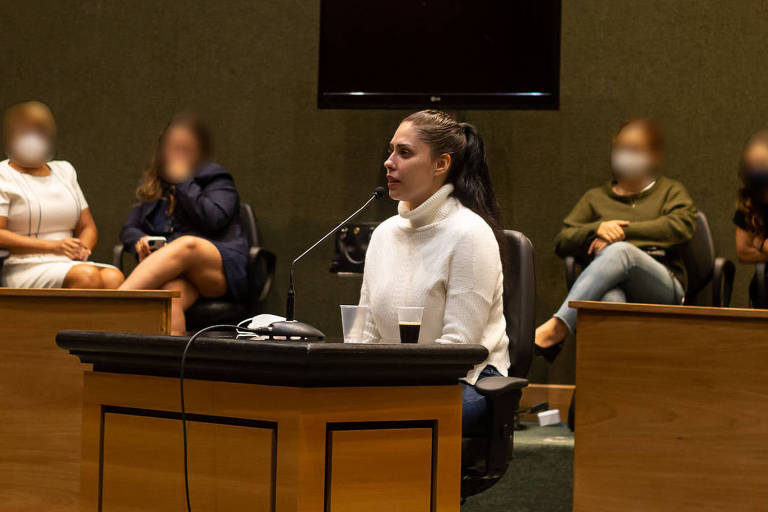 mulher de camisa branca fala ao microfone em plenário de madeira do tribunal de justiça no Rio, com público ao fundo
