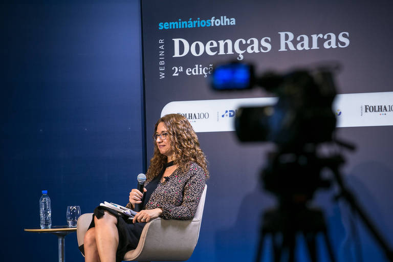 Imagem mostra Cláudia Collucci, repórter da Folha, sentada em uma cadeira branca. Na frente dela, há uma câmera e, atrás, um painel com o nome do seminário.
