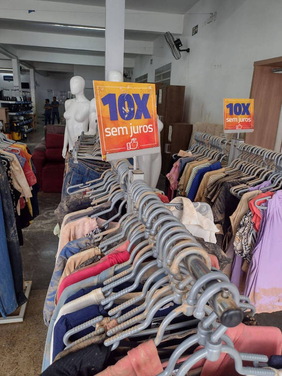 A foto mostra roupas manchadas de lama pendurada em cabides em loja do Grupo Zema em Brumadinho (MG)