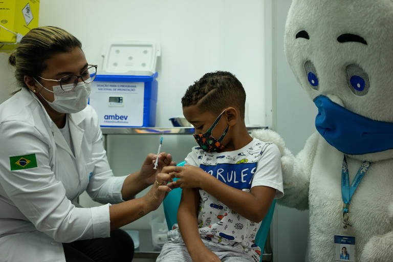 Agente de saúde de máscara e jaleco usa seringa para vacinar menino de máscara; ao lado está mascote Zé Gotinha, de máscara