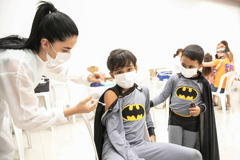agente de saúde de máscara e roupa branca aplica injeção em menino vestido de batman, acompanhado do irmão também de Batman
