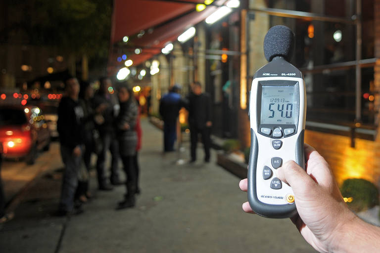 Mão segura aparelho medidor de ruídos em frente a frequentadores de bar que estão na calçada