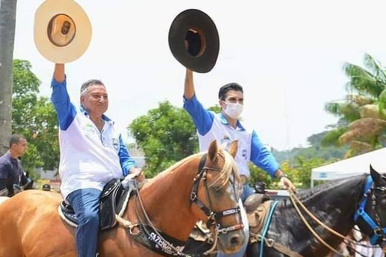 Dois homens estão montados em cavalos, um marrom e um preto; os dois usam blusas azuis e coletes brancos; eles erguem os braços e seguram chapéus em espécie de saudação