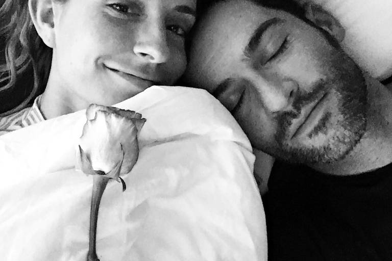 Casal posando para selfie deitados na cama, enquanto mulher segura uma rosa
