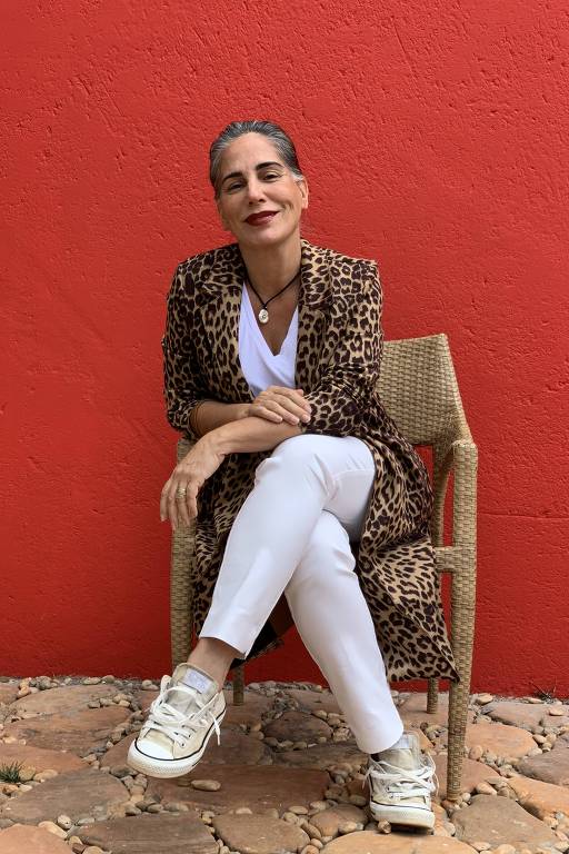 Gloria Pires está sentada em uma cadeira; ela usa calça, camiseta e tênis branco e um casaco com estampa de onça.