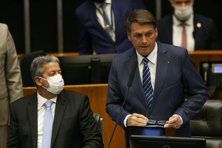 Bolsonaro, um homem branco, sem máscara, usando terno, está em pé atrás de uma mesa com notebook, com uma poltrona ao fundo e outros homens. Ao lado dele, sentado, está Lira, um homem branco, de terno e usando uma máscara, enquanto olha em direção ao presidente