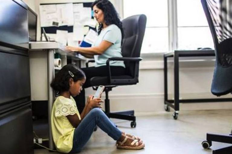 Dentro de um escritório, uma criança está sentada no chão com um celular na mão. Ao fundo, uma mulher está sentada em uma cadeira de frente para uma mesa