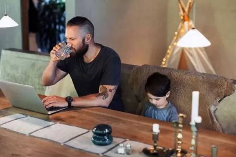 Imagem em primeiro plano mostra um homem sentado de frente para uma mesa. Ele leva um copo à boca enquanto mexe em um laptop com a outra mão. Ao seu lado, há uma criança sentada.