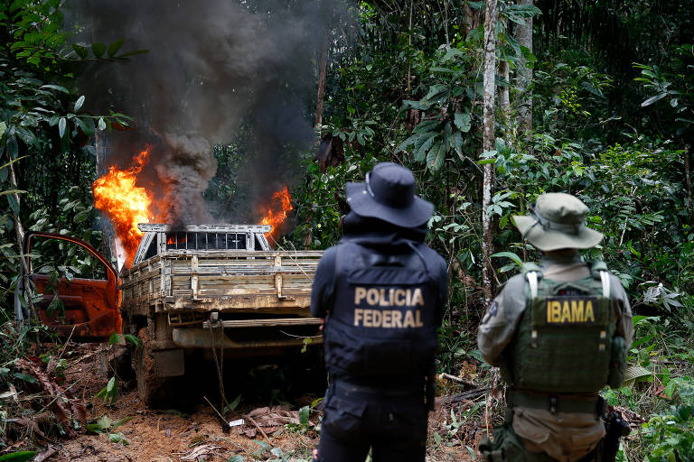 A imagem mostra um veículo em chamas em uma área de floresta densa. O fogo e a fumaça saem do carro, que está parcialmente queimado. Dois agentes, um com colete da Polícia Federal e outro com colete do IBAMA, observam a cena de costas. A vegetação ao redor é verde e exuberante, típica de uma floresta tropical.