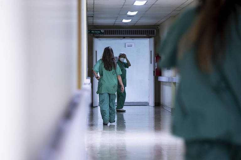 Duas mulheres se cruzam, paramentadas, em corredor de hospital