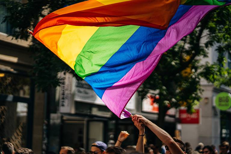 Foto mostra uma pessoa segurando acima da cabeça uma bandeira LGBTQIA+ 