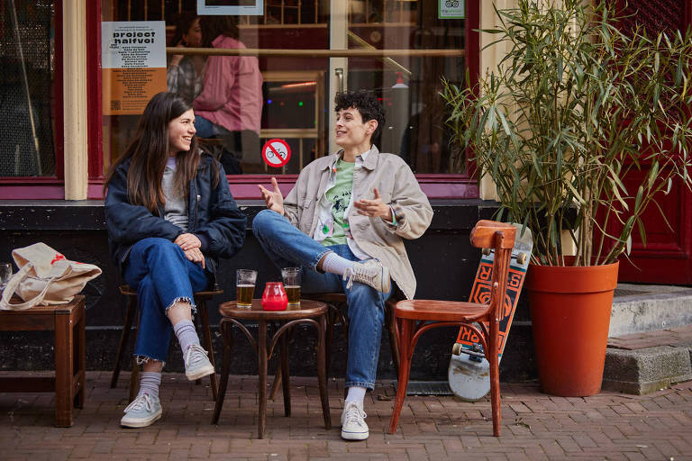 Cena de filme em que dois jovens estão sentados em bancos numa calçada conversando e tomando cerveja
