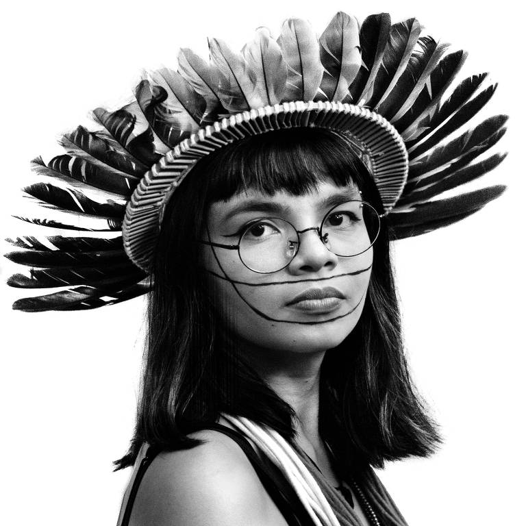 retrato em preto e branco de Txai, jovem mulher indígena. ela está de perfil, olhando para a câmera, e tem cabelos pretos e lisos que vão até os ombros. ela usa óculos de aros redondos e finos. na cabeça, veste cocar. 