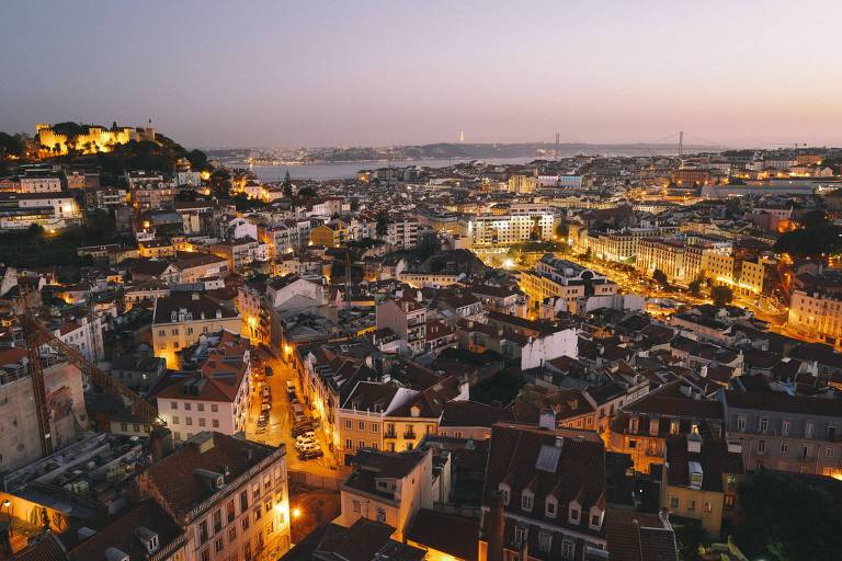 Foto mostra vista aérea de Lisboa, com prédios cortados por ruas, onde a iluminação amarelada se destaca. No fundo, é possível ver a ponte 25 de Abril, que corta o Rio Tejo. A imagem foi tirada no fim da tarde, por isso o céu está escurecendo.