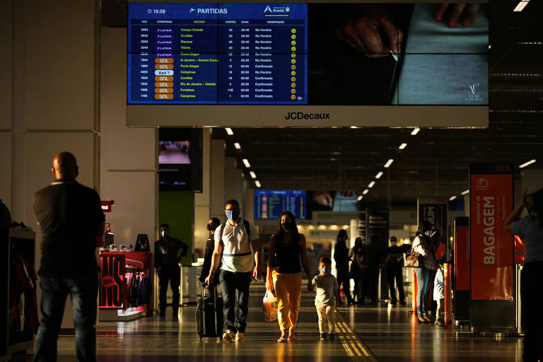 Pessoas andando com malas no Aeroporto Internacional de Brasília (DF). Painel de voos na parte superior da foto.