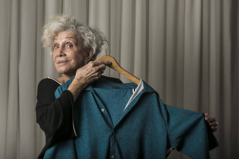 Foto mostra Helena Schargel, 82, mulher de cabelos grisalhos, que usa roupa preta e segura uma peça de roupa verde escura.