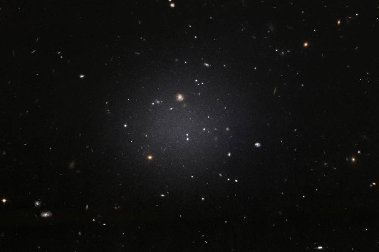 Uma galáxia tênue, desprovida de gás, em foto do telescópio Hubble, em que é possível ver outras galáxias de fundo através dela