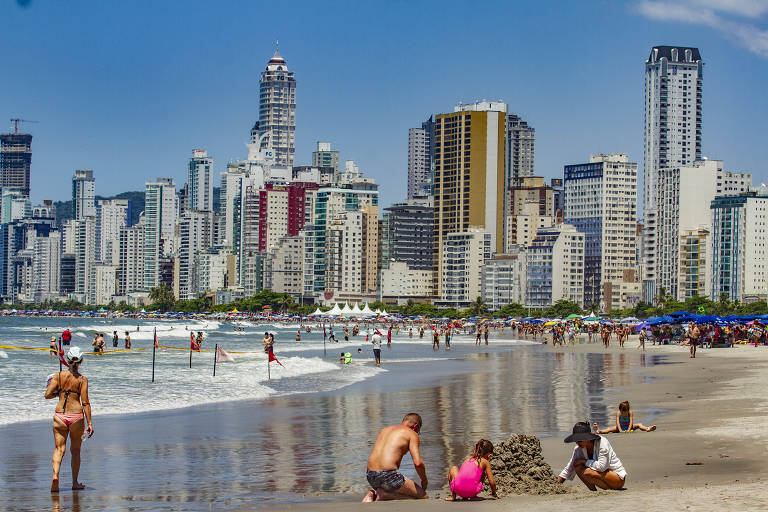 Crianças fazendo monte de areia em praia com muitas pessoas e prédios altos ao fundo, em dia de sol 