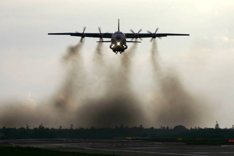 Um avião grande paira no ar; das turbinas, saem muitas fumaça marrom acinzentada