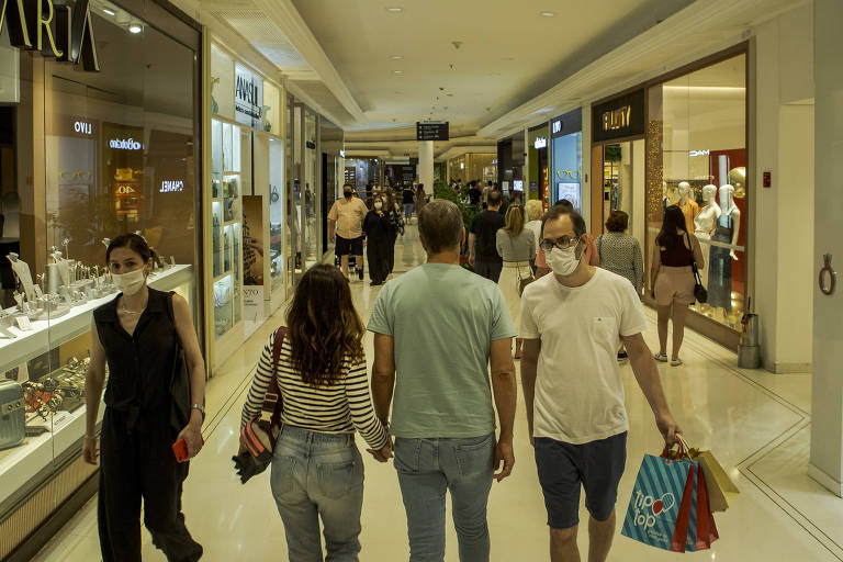 Imagem mostra pessoas caminhando num corredor de um shopping. Algumas seguram sacolas de compras.