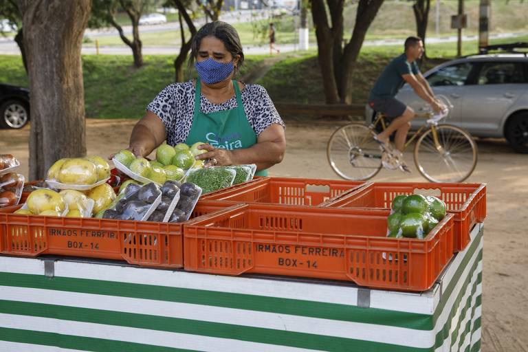 Mulher espalha frutas sobre uma mesa enquanto ciclista passa ao fundo