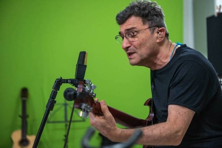 Em foto colorida, o compositor e instrumentista Hélio Ziskind aparece cantando e tocando violão