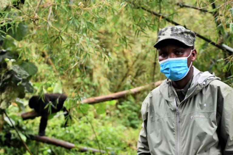 Homem negro de máscara posa para foto. Ao fundo, se vê um gorila pendurada em um galho de árvore.