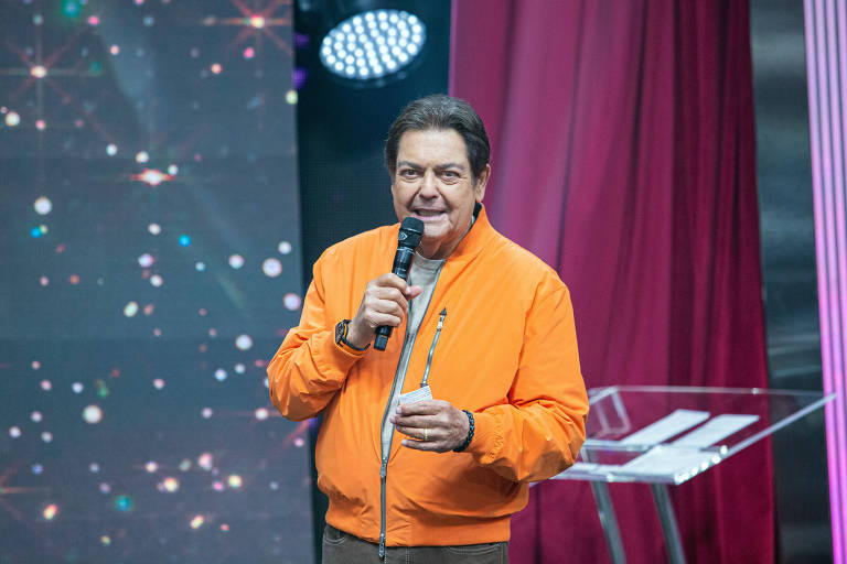 Fausto Silva de jaqueta cor de laranja, com um microfone na mão e um papel na outra; ele está no palco, mas imagem só o mostra da cintura para cima, em plano americano 