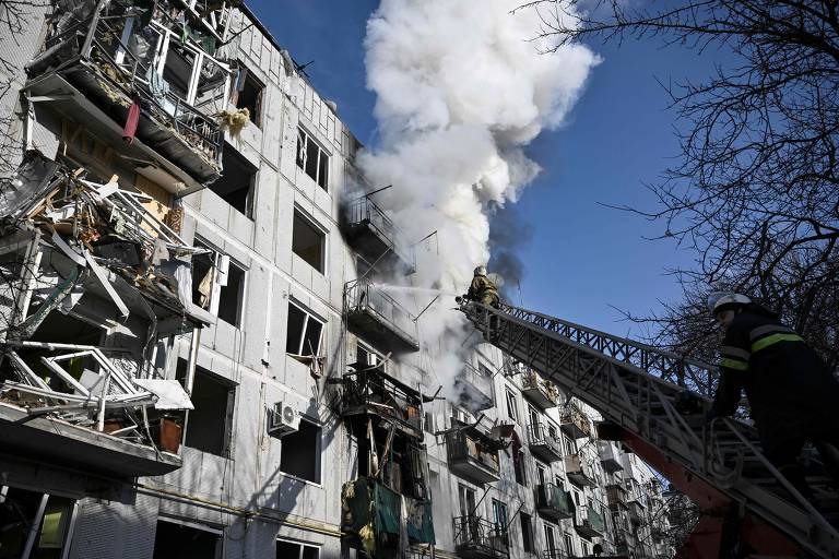 bombeiros em escada diante de edifícios em chamas - vê-se muita fumaça e, ao fundo, o céu muito azul