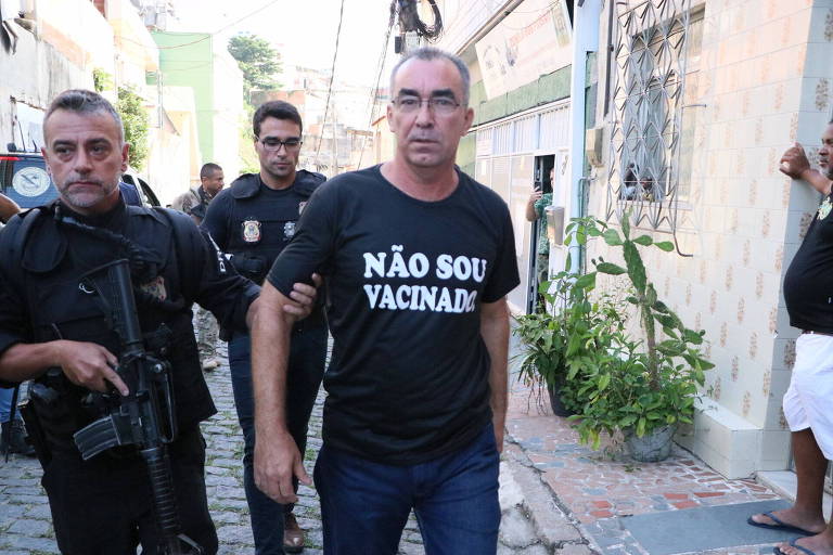 homem com camisa preta escrita NÃO SOU VACINADO é conduzido por policial de fuzil na rua, com outros policiais atrás