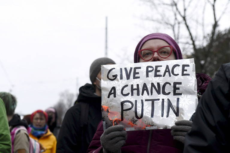 Mulher de gorro de inverno cor de vinho e óculos na mesma cor segura cartaz com os dizeres "Dê uma chance à paz Putin", em inglês. O cartaz cobre seu rosto do nariz para baixo
