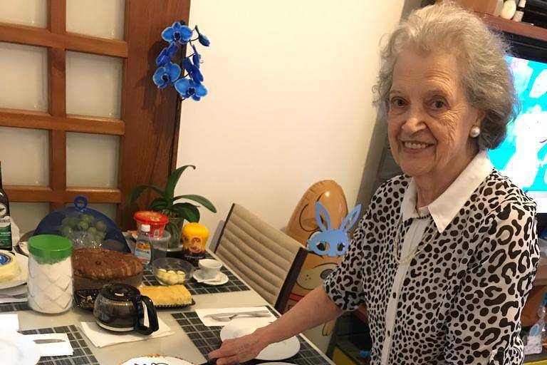 Imagem em primeiro plano mostra mulher idosa sorrindo e posando para foto ao lado de uma mesa com um bolo de aniversário
