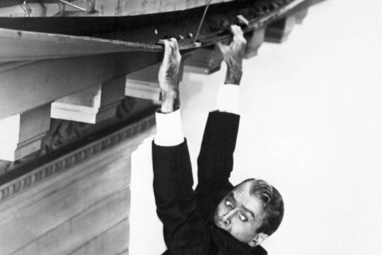 Um homem de terno está pendurado da calha de um telhado com um olhar apavorado. Imagem em preto e branco