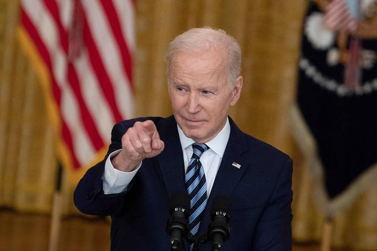 Biden, de terno escuro e gravata listrada azul, aponta o indicador direito para a câmara