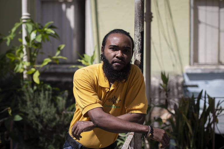 Homem negro, de barba, usando um camisa amarela está com os braços cruzados, apoiando-se em um portão branco