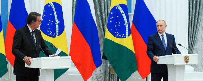 Vladimir Putin e Jair Bolsonaro em pé, de terno, flam em púlpito branco com as bandeiras da Rússia e Brasil ao fundo