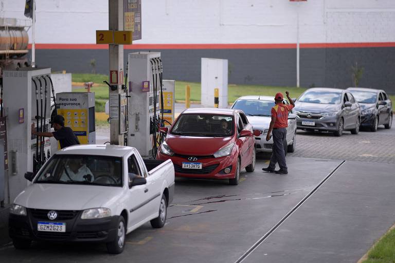 Carros aguardam em fila para serem abastecidos em posto de gasolina. É possível ver um frentista acenando para organizar os motoristas