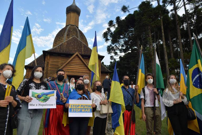 Manifestantes seguram bandeiras da Ucrânia e cartazes de protesto em parque, com céu azul
