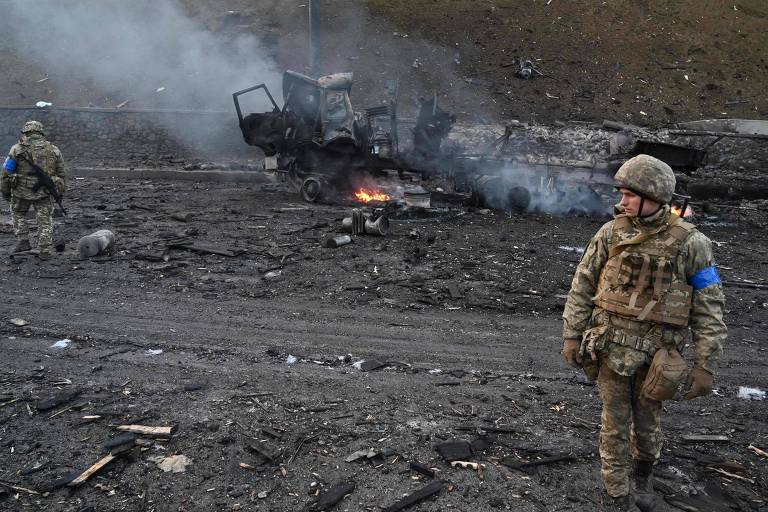 Soldado fardado olha para o lado em cenário de destruição, com outro soldado de costas e veículo carbonizado soltando fumaça ao fundo