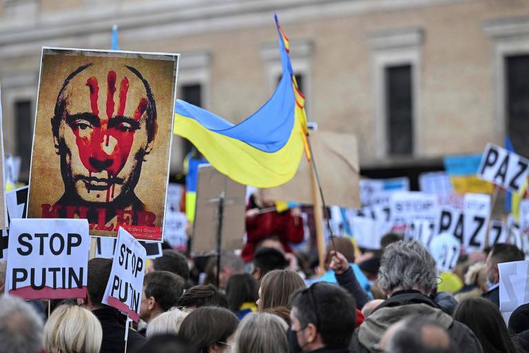 Um grupo de pessoas aglomeradas com bandeiras da Ucrância e cartazes com os dizeres "paz", "stop putin" e uma placa com o rosto de Putin com a marca de uma mão sangrando