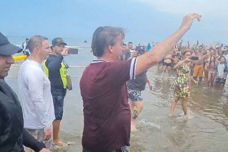 Bolsonaro, um homem branco, camiseta vinho, na praia cumprimentando e tirando fotos com pessoas que se aglomeram atrás de uma grade de metal