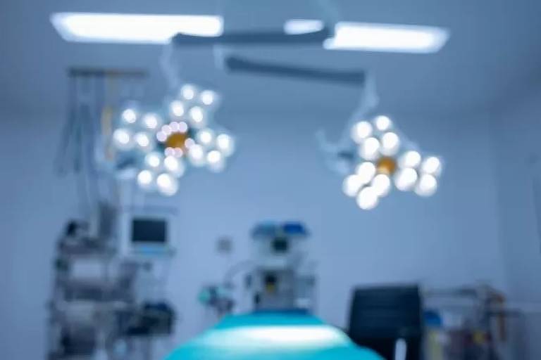 Imagem desfocada mostra uma sala de cirurgia com maca e equipamentos médicos