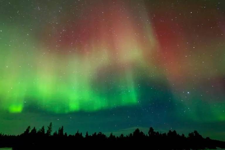 Imagem panorâmica mostra aurora boreal com céu em tom de verde, azul e vermelho