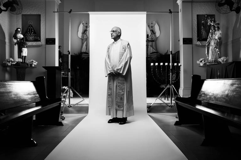 Vestindo uma túnica, Padre Julio posa em frente a um fundo branco montado no salão de uma igreja