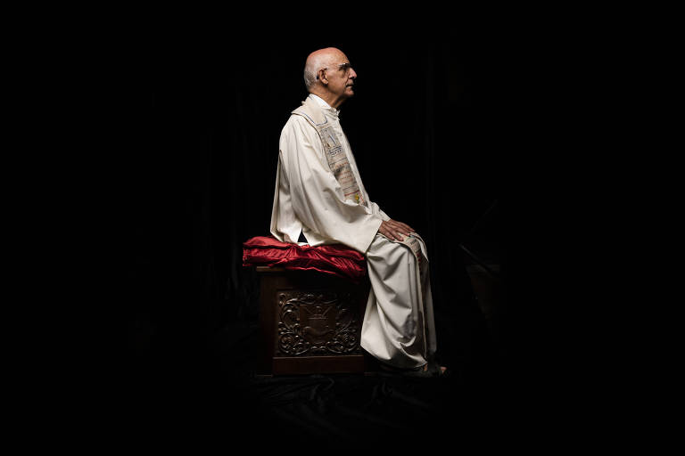 Vestindo uma túnica, Padre Julio posa sentado, de perfil, em frente a um fundo preto