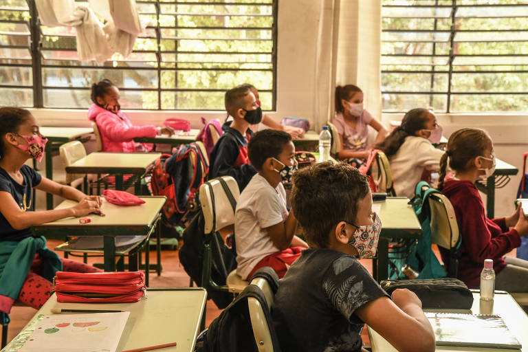 Várias crianças sentadas em suas carteiras numa sala de aula, vestidas com roupas coloridas e usando máscaras de proteção contra o coronavírus. A sala é bem iluminada, com janelas amplas que permitem observar a vegetação na área externa da escola.