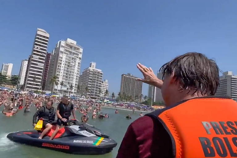 De colete laranja com os dizeres "presidente Bolsonaro" nas costas, Bolsonaro acena para banhistas que estão dentro do mar. Vários prédios aparecem ao fundo e na frente dos banhistas há dois homens em um jet-ski