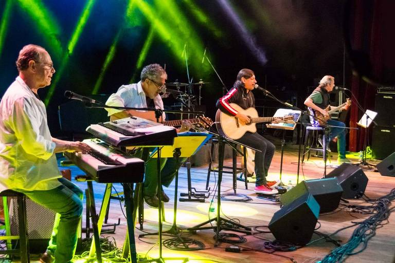 O grupo 14 Bis aparece, em foto colorida, cantando e tocando em um palco