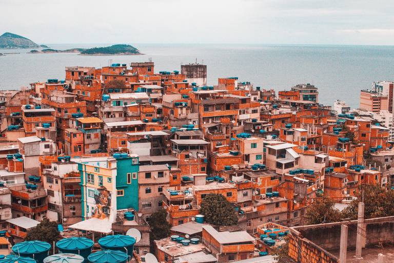 Foto mostra um amontoado de casas, com tijolos à vista, formando a favela do Cantagalo. No fundo, é possível ver o oceano