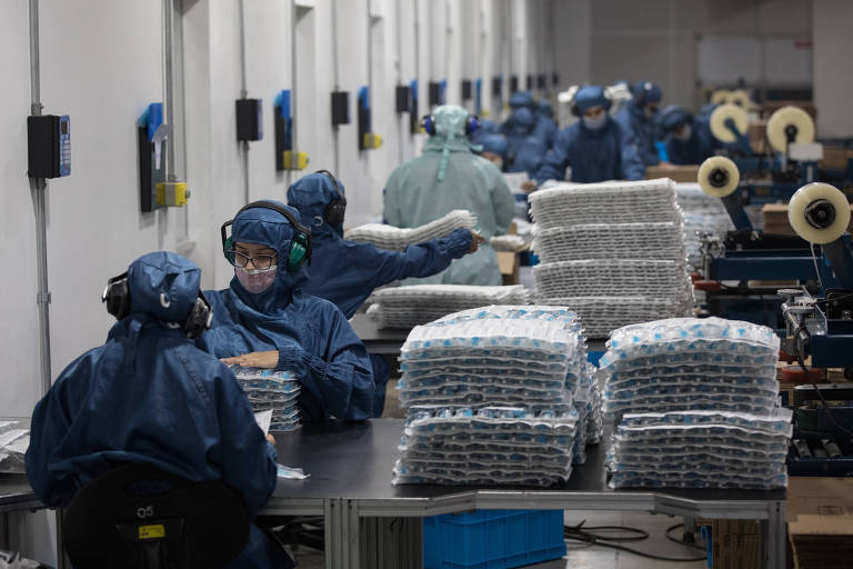 Pessoas de macacão azul e máscara trabalhando em mesa, em fábrica 